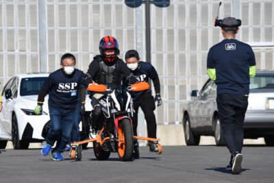 転倒を防止するためのアウトリガーを装着した小型バイクに乗車し、まずはエンジンを掛けずにボランティアスタッフの手押しから走行を始める