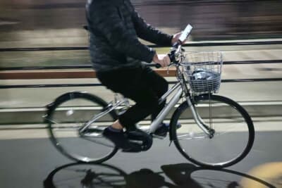 夜間、シティサイクルを片手運転しながらスマートフォン操作。画面を見つめる視線は前方から逸れており、重大な事故を招く危険な行為だ。速やかにやめるべきである
