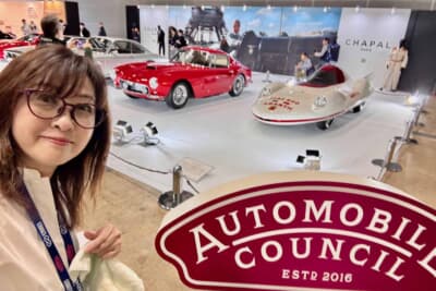 吉田由美さんがオートモビル カウンシルのロゴの前で一枚。後方には主催者展示「Designed by ピニンファリーナ」コーナーの「フェラーリ 250GT SWB」（赤）と「フィアット アバルト 750 レコルド エンデューロ ピニンファリーナ」（白）が並ぶ