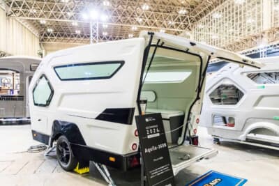 ダイレクトカーズ Aquila360N:ジャパンキャンピングカーショー2026のブースに展示された、流線型ボディと幾何学的な窓が特徴の白い車体。後部ハッチを開いた状態